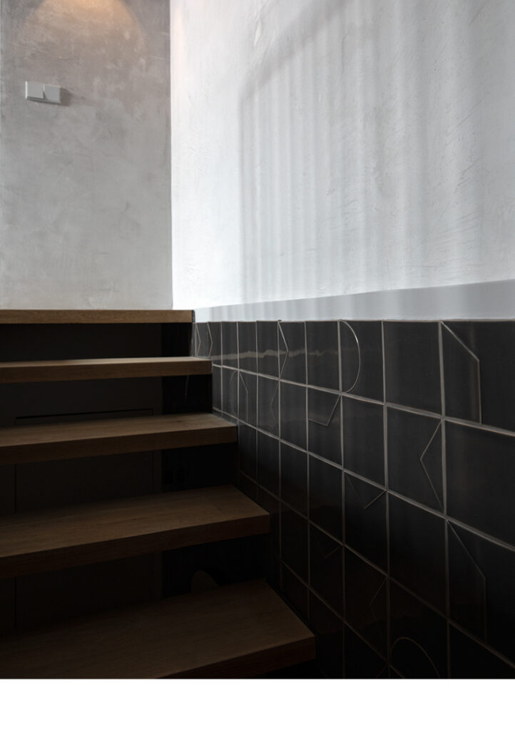 Timber stairs beside a dado of dark charcoal Smink Studio relief tiles below a white wall.