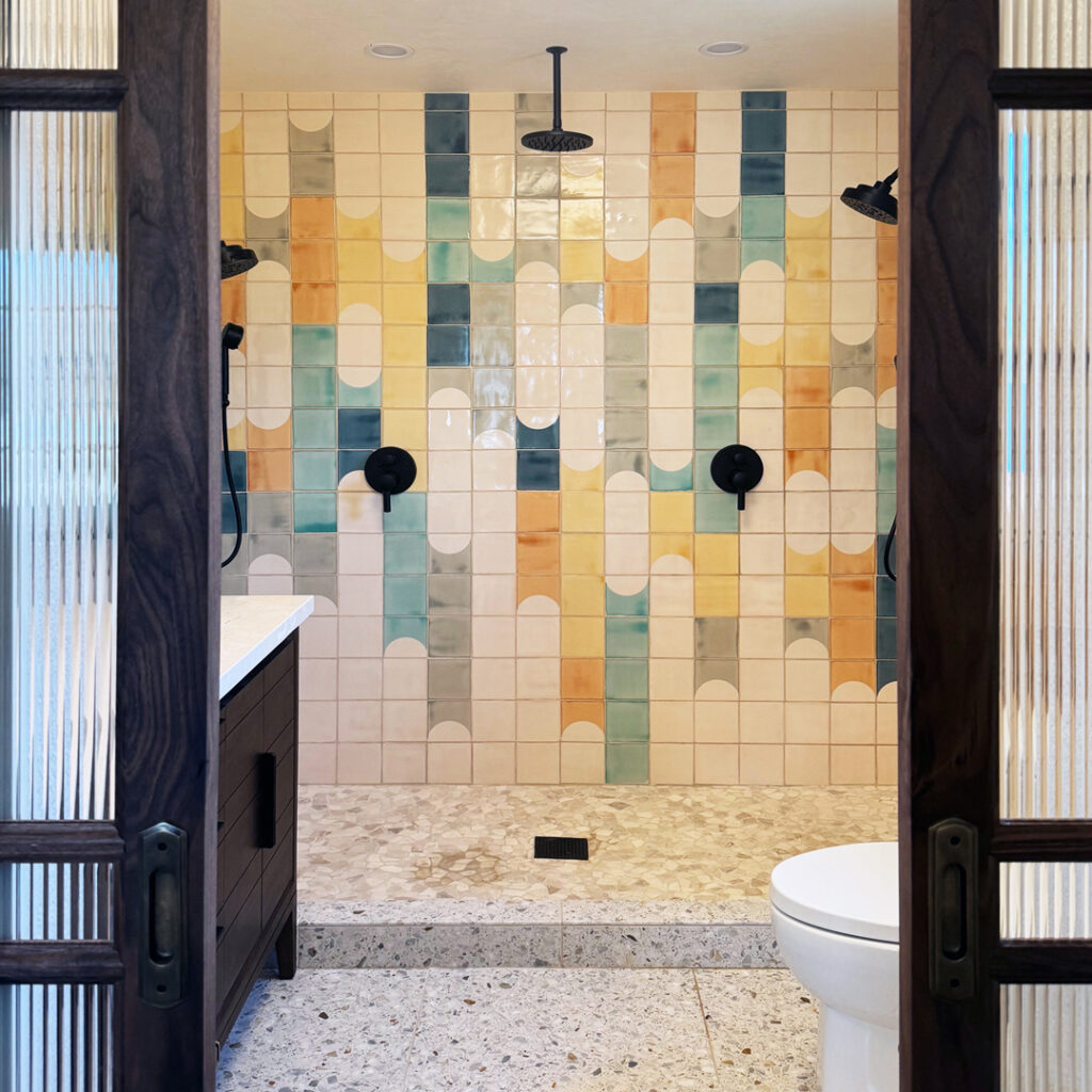 View through glass doors into a twin shower lined with Smink Studio Waterfalls glazed ceramic tiles in teal, yellow and orange.