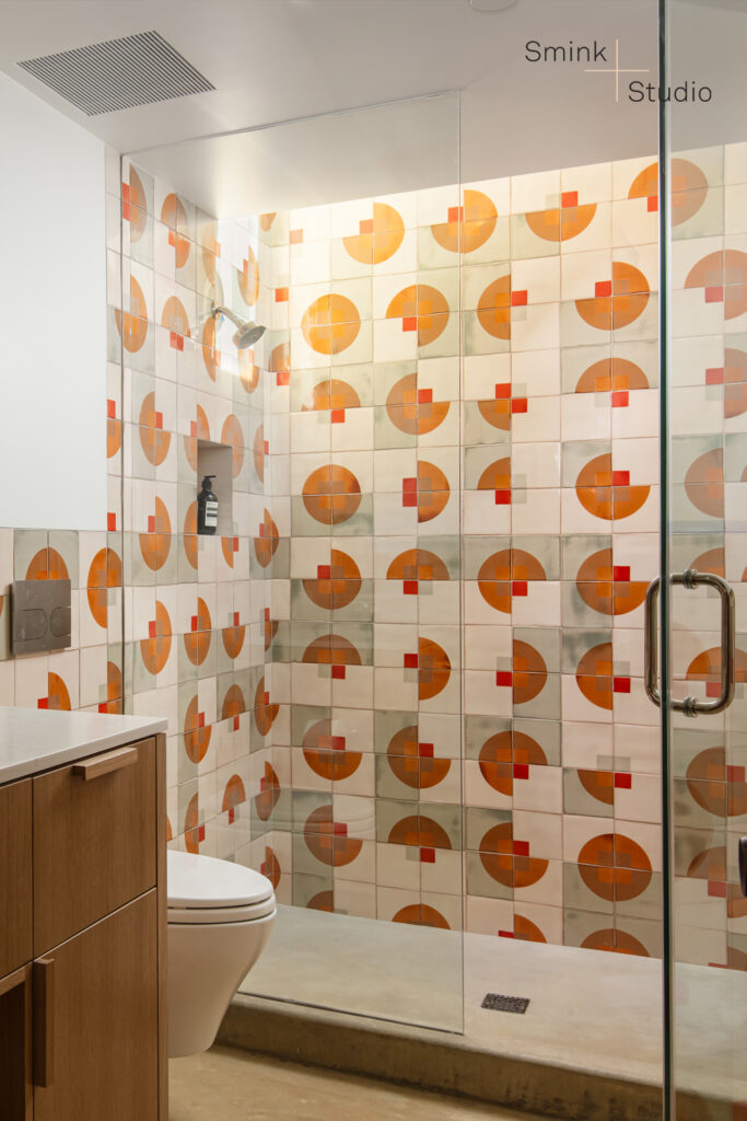 Glass shower lined with cream Smink Studio handmade tiles bearing screen-printed orange half-circles and red squares.
