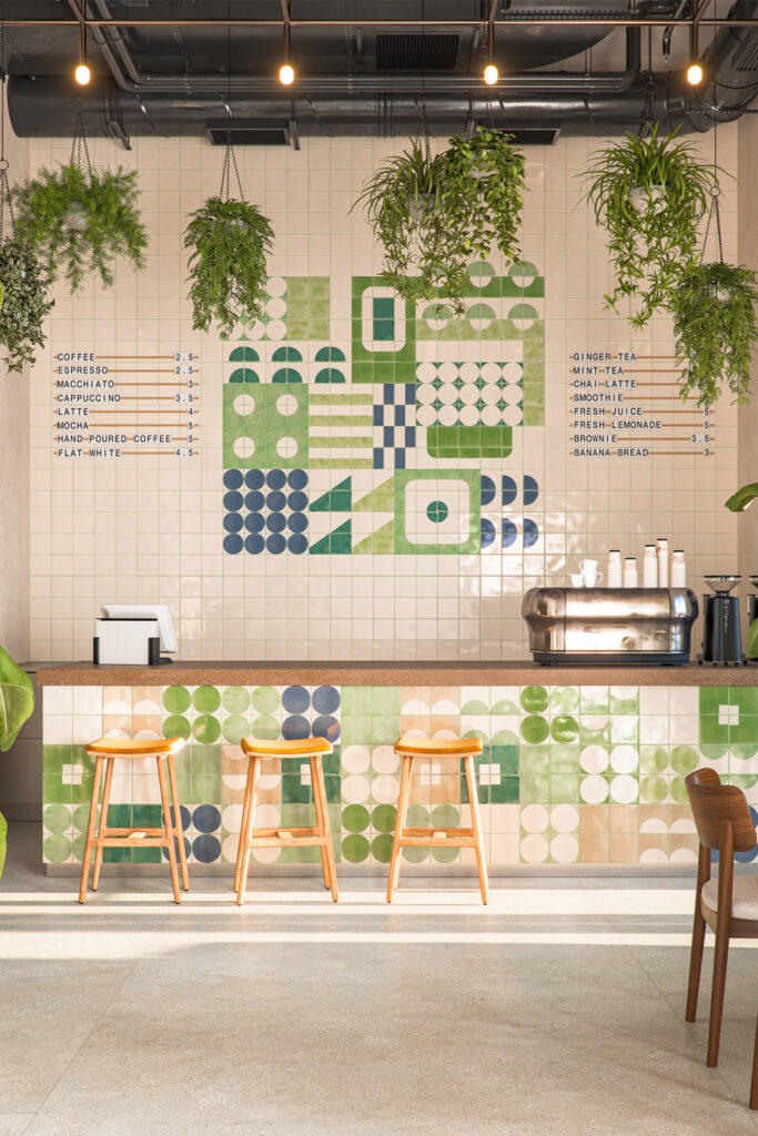 Café interior with hanging plants and a feature wall of Smink Studio Bauhaus-inspired glazed ceramic tiles in greens and navy.