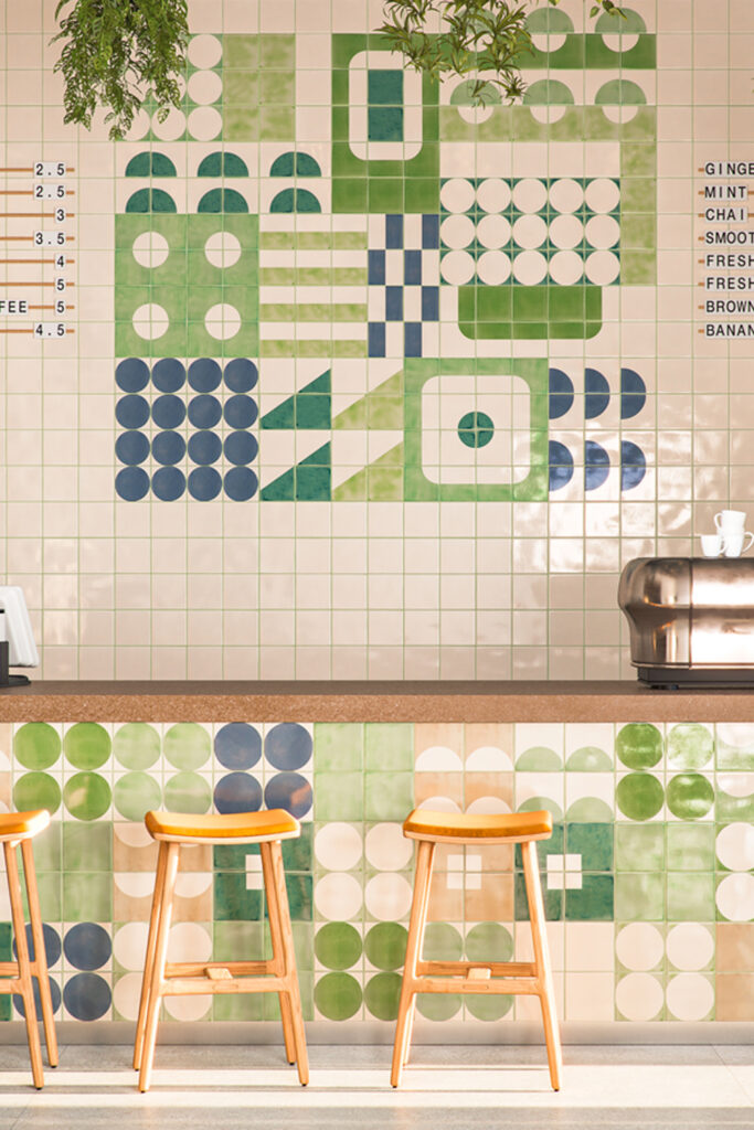 Café counter with stools before a menu wall of Smink Studio Bauhaus screen-printed tiles in green, sage and navy.