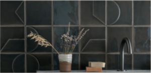 Smink Studio relief tiles in a dark charcoal glaze with raised line patterns, behind a kitchen tap and dried flowers.