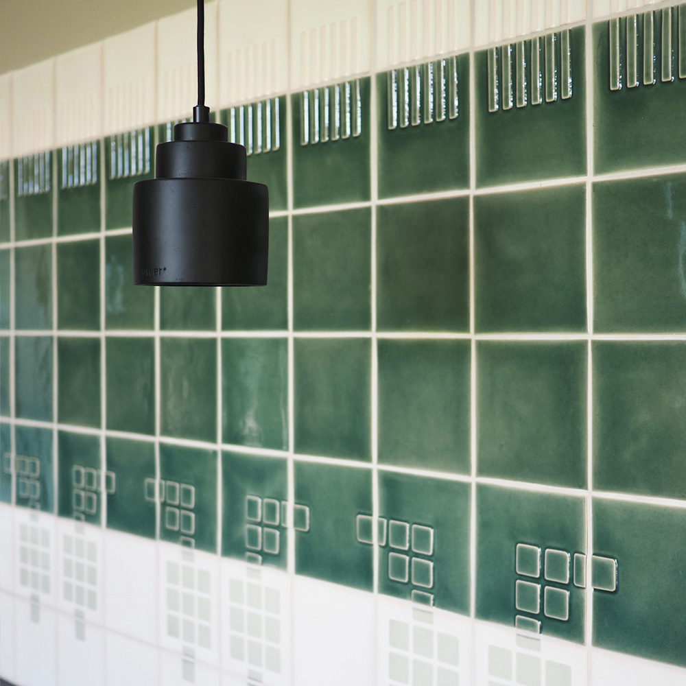 Wall of dark green Smink Studio glazed ceramic tiles above a white band of screen-printed squares, with a black pendant lamp.