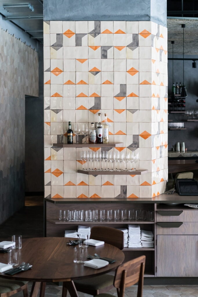 Restaurant bar column clad in Smink Studio handmade ceramic tiles with a cream, orange and grey geometric pattern in screen-printed colours, with shelves of wine glasses and bottles.