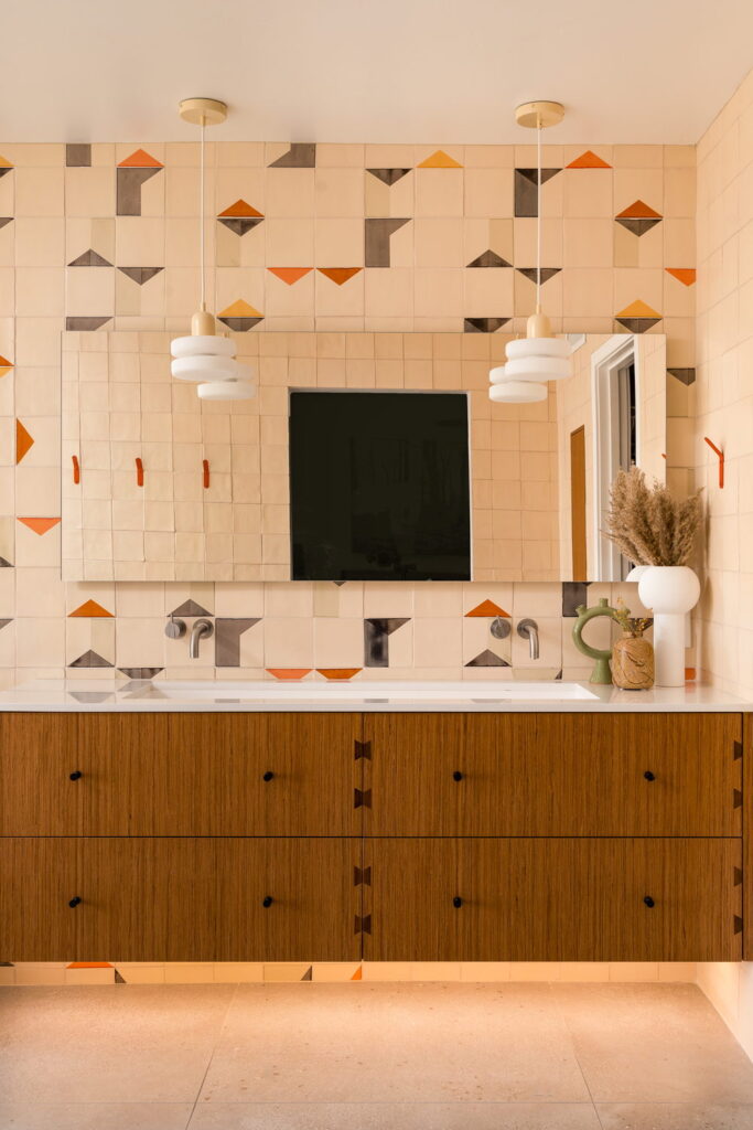 Bathroom vanity backed by Smink Studio handmade ceramic tiles in cream with orange, mustard and charcoal geometric screen-printed colours, with an oak vanity and twin pendant lights.