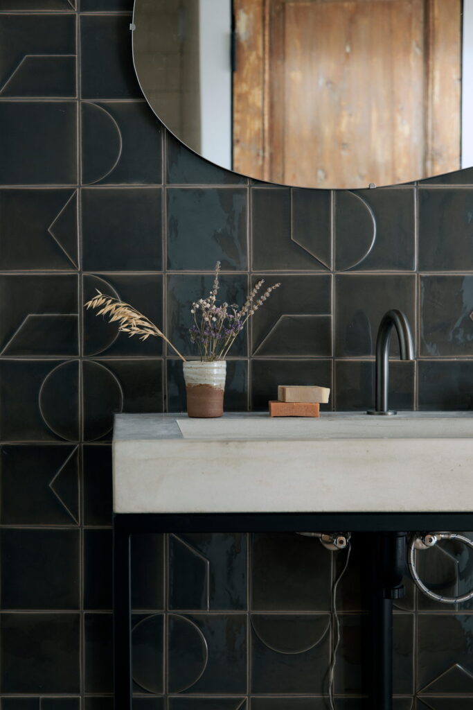 Bathroom of glossy near-black Smink Studio handmade ceramic tiles with tonal geometric screen-printed colours, with a concrete basin, round mirror and black tap.