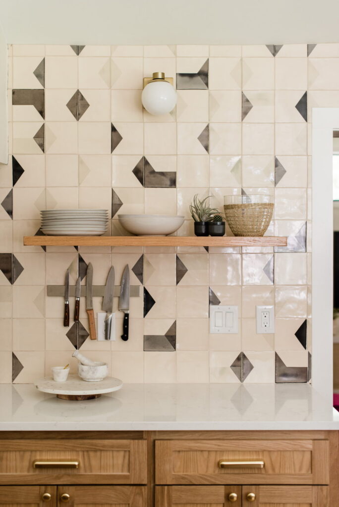 Kitchen splashback detail of Smink Studio handmade ceramic tiles in cream with charcoal diamond and geometric screen-printed colours, with an oak shelf and brass sconce.