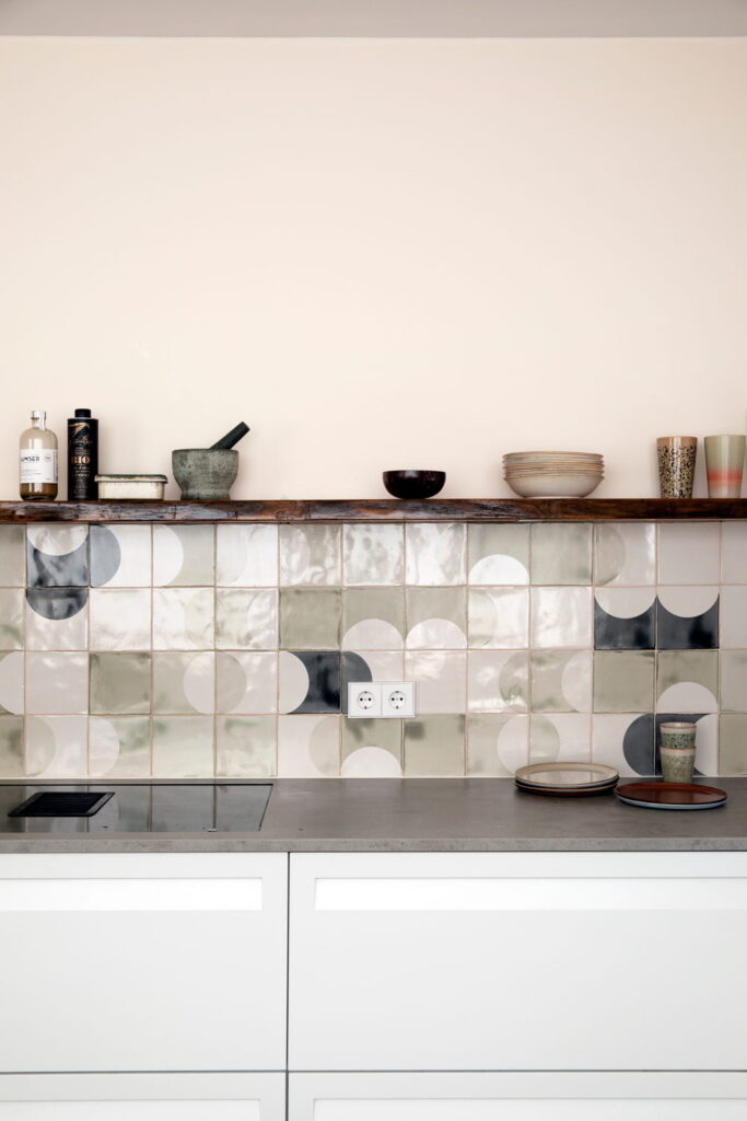 Kitchen splashback of Smink Studio handmade ceramic tiles in sage-green, grey and cream with half-circle screen-printed colours, beneath a rustic wood shelf.