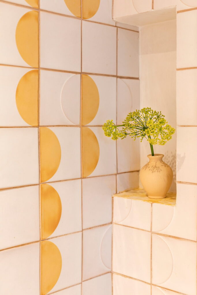 Shower niche of white Smink Studio handmade ceramic tiles with mustard-yellow half-circle screen-printed colours, styled with a small vase of flowers.