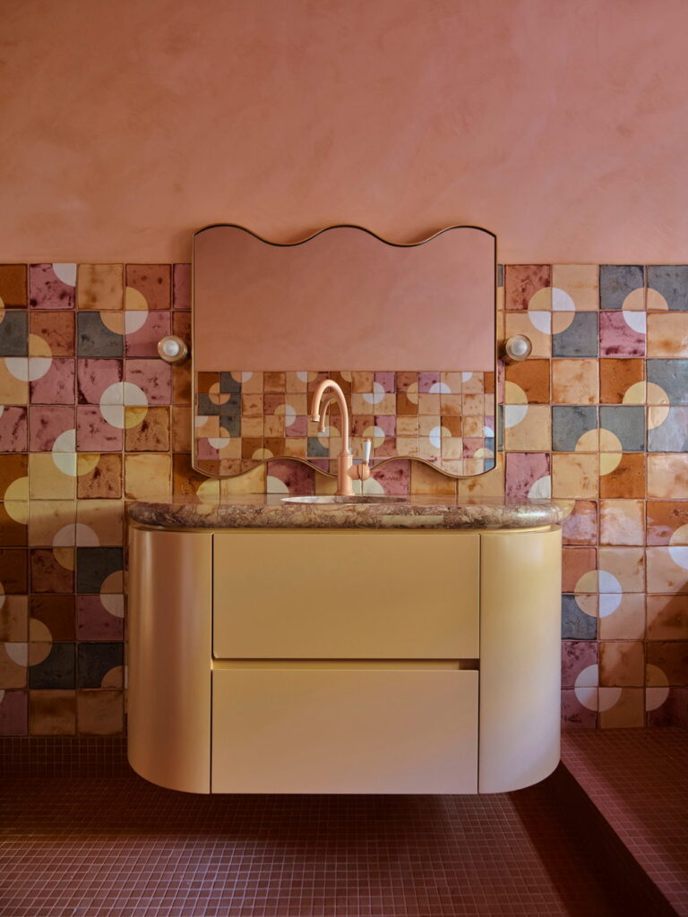 Bathroom vanity backed by multicoloured Smink Studio handmade ceramic tiles in pink, blue, ochre and terracotta circle screen-printed colours, with a wavy mirror and curved vanity.