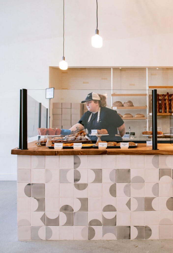Bakery counter clad in white and grey Smink Studio handmade ceramic tiles with half-circle screen-printed colours, with breads and pastries behind the glass.