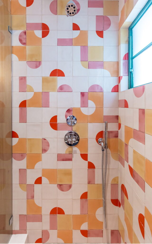 Shower of cream Smink Studio handmade ceramic tiles with pink, coral, orange and mustard half-circle screen-printed colours, with chrome fittings and a turquoise-framed window.