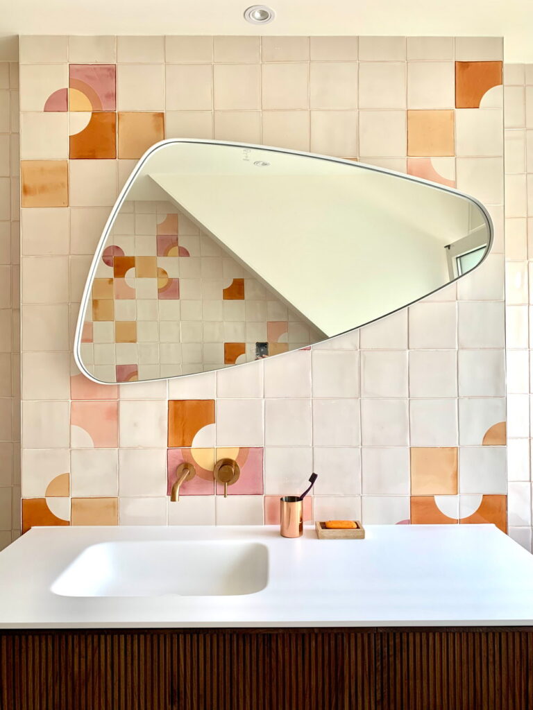 Bathroom vanity backed by cream Smink Studio handmade ceramic tiles with pink, apricot and mustard quarter-circle screen-printed colours, with an organic mirror and fluted vanity.