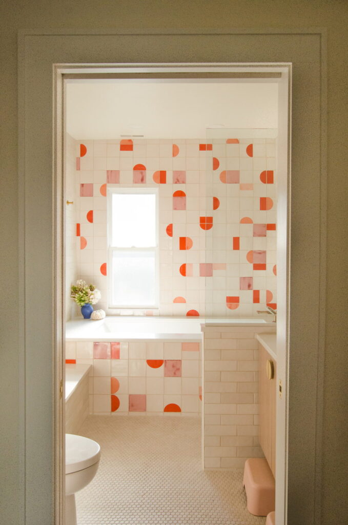 Bathroom of white Smink Studio handmade ceramic tiles with coral-red and pink half-circle screen-printed colours, framing a bath beneath a window.