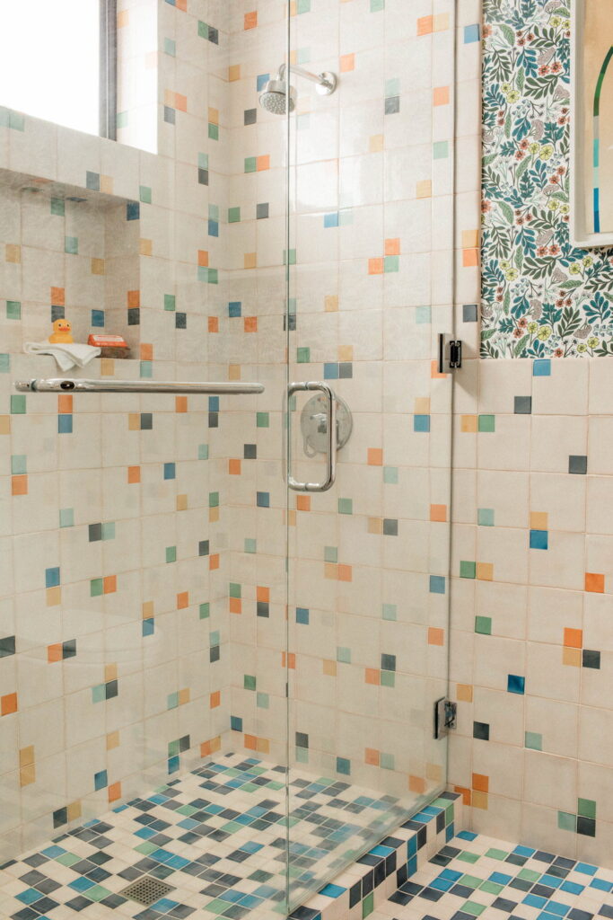 Shower of cream Smink Studio handmade ceramic tiles scattered with orange, blue, green and yellow square accents in screen-printed colours, behind a glass screen.