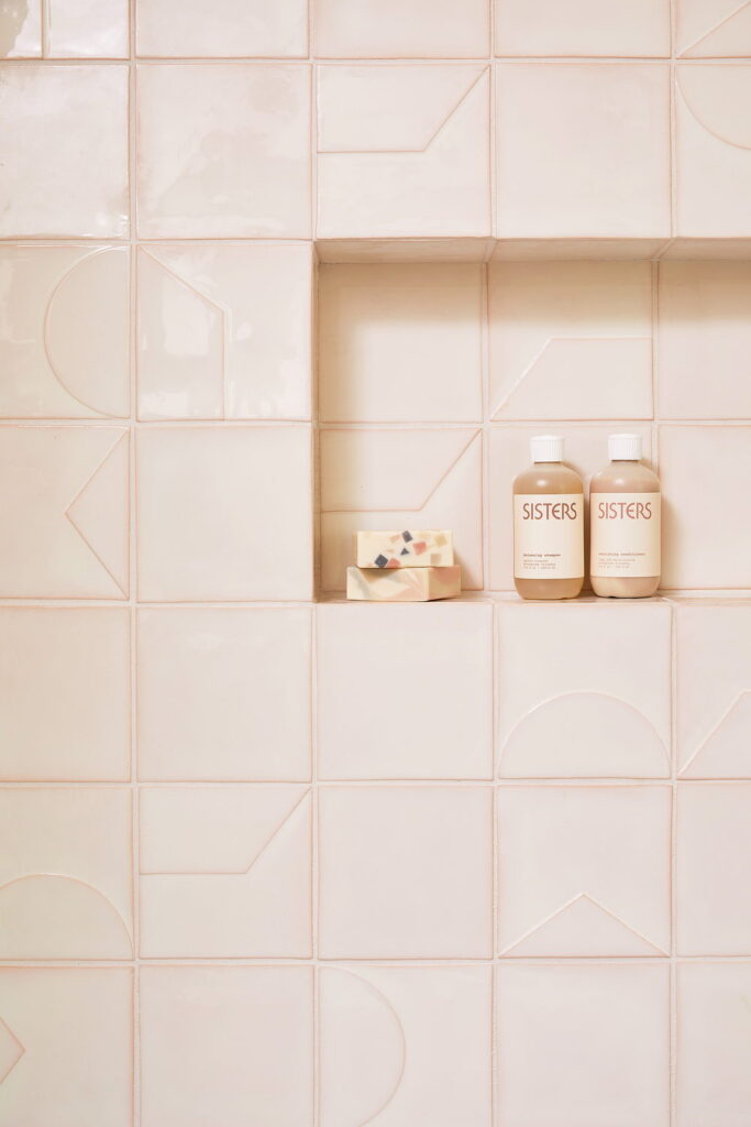 Shower niche of blush-pink Smink Studio handmade ceramic tiles with embossed geometric line detailing, styled with soap and bottles.