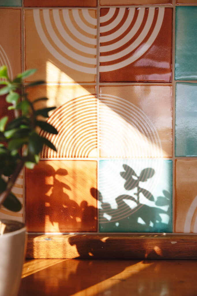 Smink Studio handmade ceramic tiles in amber, terracotta, teal and green with a concentric-circle relief pattern, lit by sunlight and leaf shadows.