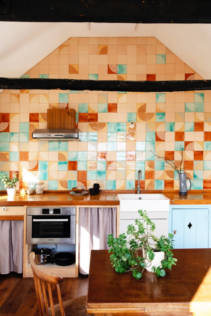Kitchen with a full wall of Smink Studio handmade ceramic tiles in caramel, teal and terracotta with concentric-circle relief patterns, beneath a beamed ceiling.