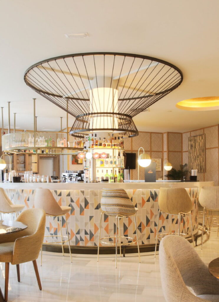 Hotel bar with a curved front of Smink Studio handmade ceramic tiles in cream, orange and grey triangle screen-printed colours, with upholstered stools and a wire pendant.
