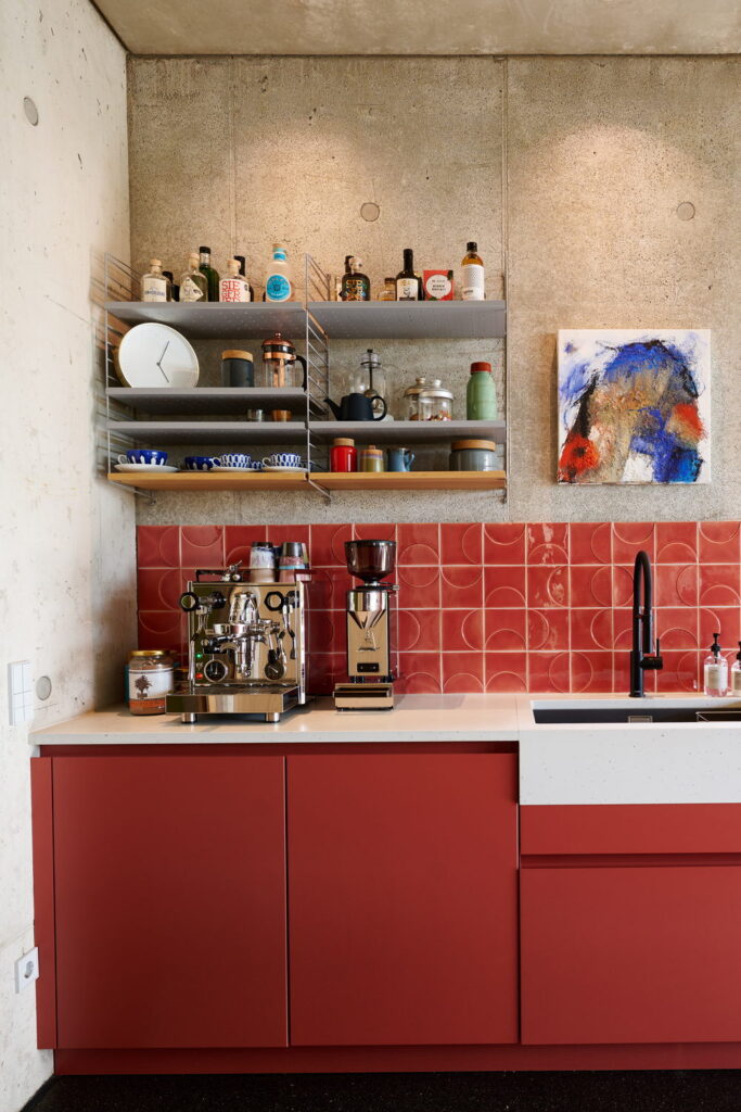 Coffee corner backed by red Smink Studio handmade ceramic tiles with half-circle screen-printed colours, against a concrete wall with an espresso machine and red cabinets.
