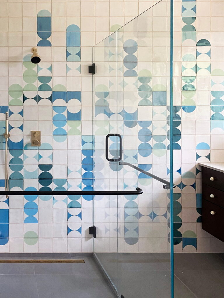 Shower of Smink Studio handmade ceramic tiles in blue, green, teal and cream half-circle screen-printed colours, behind a glass screen with black and brass fittings.