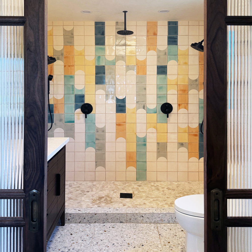 Double shower of Smink Studio handmade ceramic tiles in cream, yellow, teal and grey half-circle screen-printed colours, with twin black rain showers and a terrazzo floor.