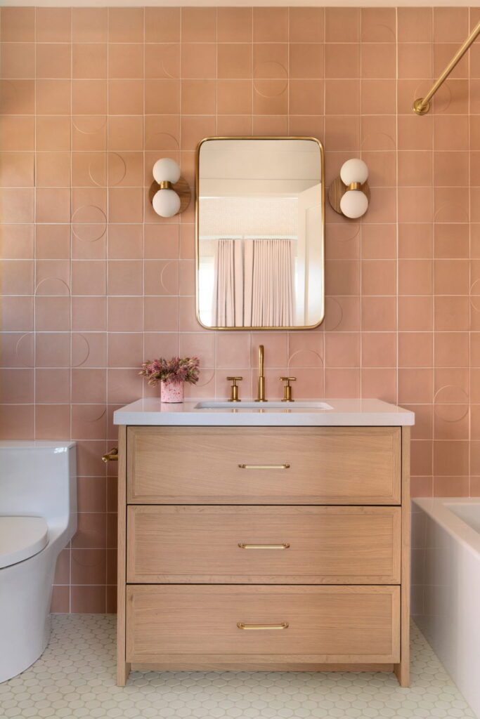 Bathroom vanity backed by terracotta and peach Smink Studio handmade ceramic tiles with subtle embossed circle detailing, brass mirror and sconces.