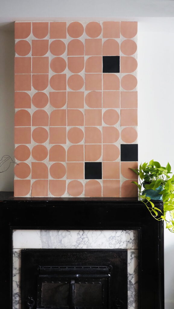 Fireplace panel of unglazed terracotta Smink Studio handmade ceramic tiles in circle, half-circle and arch shapes, with two black square accents, above a black-and-marble fireplace.