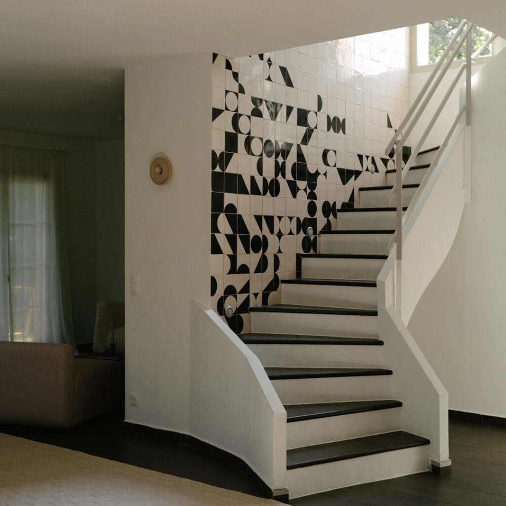 White winding staircase rising beside a wall of black and white Smink Studio screen-printed tiles in an Imraum Design home.