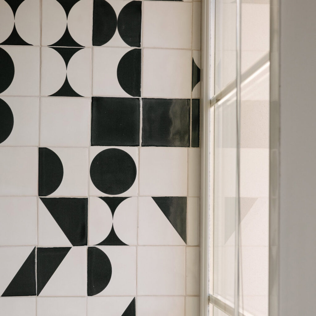 Corner of a wall in white handmade glazed ceramic tiles with black screen-printed half-circles and triangles, beside a tall window.