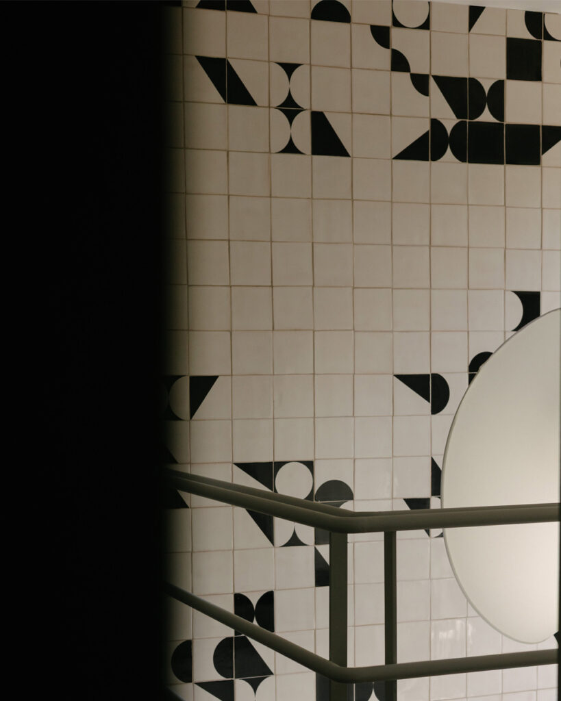 Stairwell wall of white Smink Studio handmade tiles with black screen-printed circles, triangles and half-circles, behind a steel handrail and paper lamp.