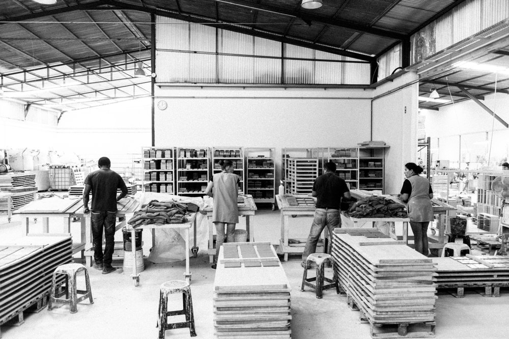 Black and white photograph of the Portuguese factory where Smink Studio tiles are made, with workers at benches and stacks of unfired tiles on pallets beneath a pitched roof.