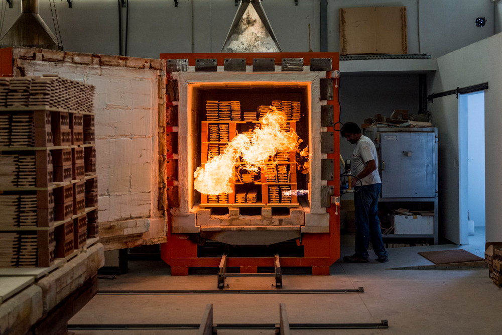 The open door of a kiln at the Portuguese factory, flames burning inside, with a loaded kiln car of Smink Studio tiles waiting alongside.