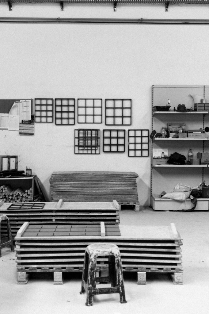 Black and white photograph of the factory workshop, with metal tile moulds hanging on the wall above stacked pressing boards on pallets.