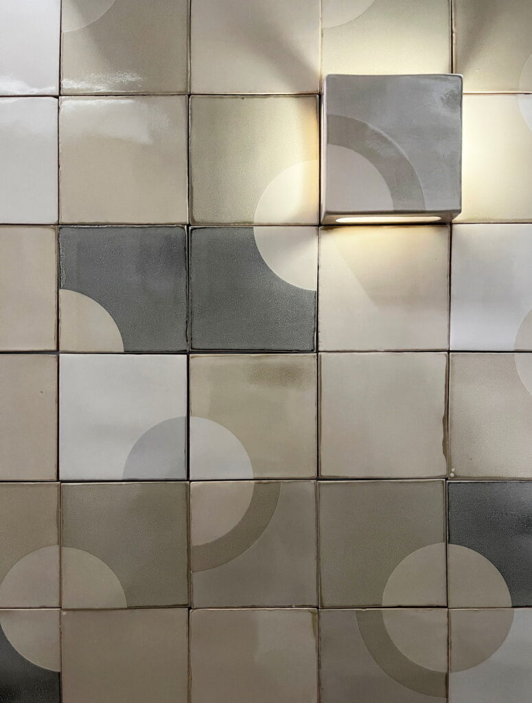 A wall of grey screen-printed Smink Studio tiles with a lit pocket tile set among them, its recess holding a bulb that casts warm light across the surrounding surface.