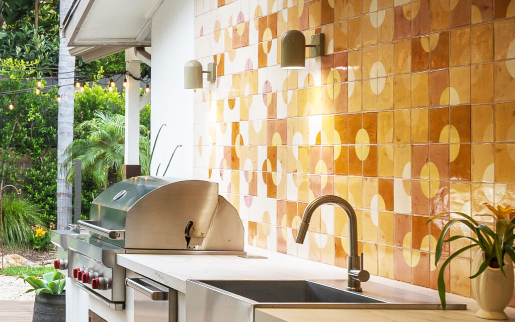 Outdoor kitchen by Brookside, with a grill and sink backed by Smink Studio tiles in honey, amber and orange with cream circles.