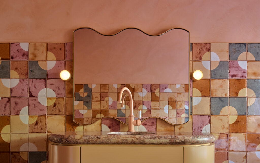 Bathroom by YSG Studio: a wavy mirror and curved vanity before Smink Studio tiles in rose, terracotta, slate and honey with white circles.